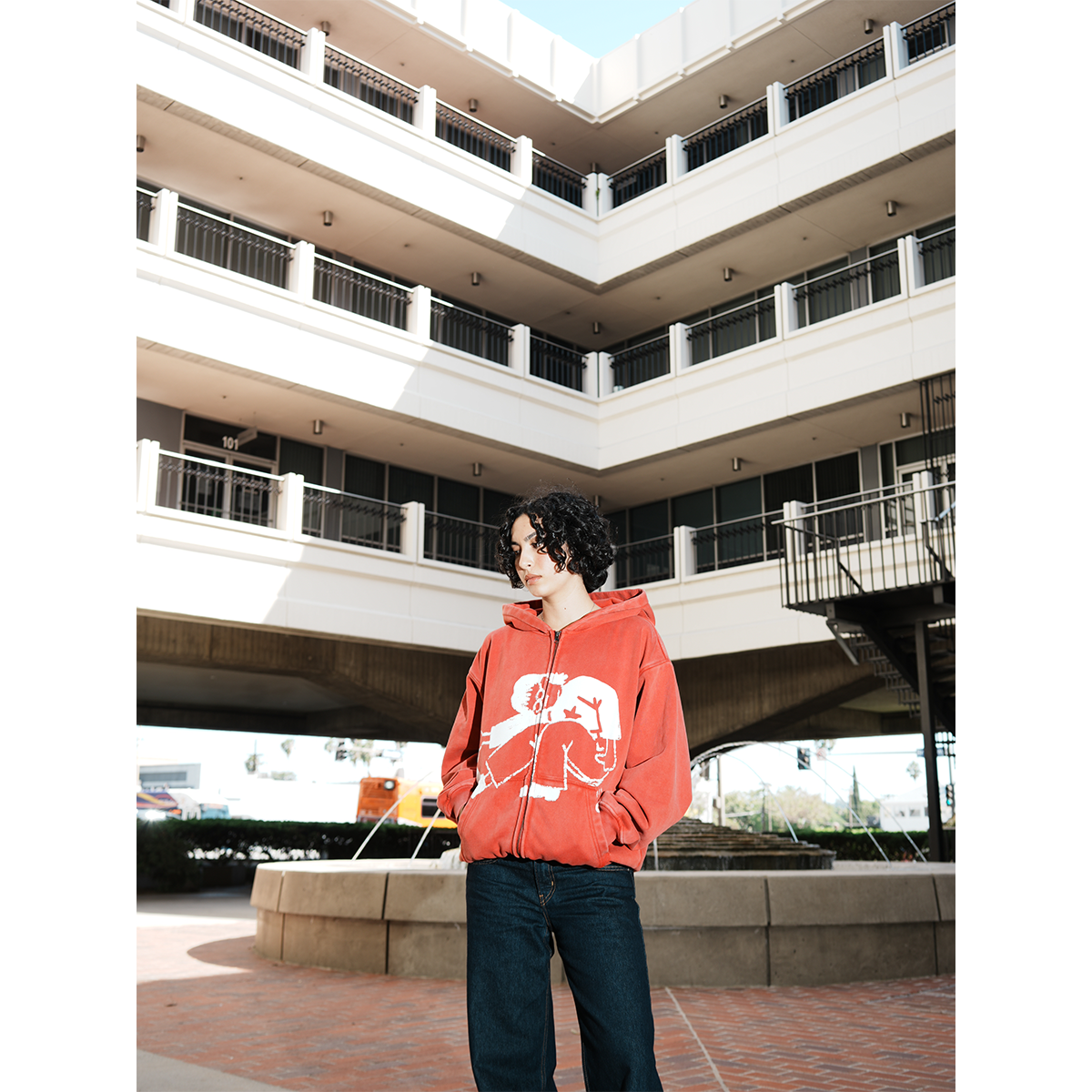 Model wearing a red hoodie with a white graphic design in an outdoor setting with hands in pocket.